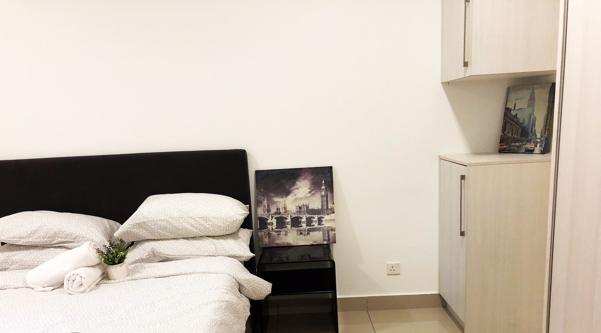 A neatly made bed with pillows and a rolled towel, a small bedside table with a plant, and a wall-mounted cabinet with a picture.