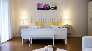 Un dormitorio con una cama blanca, dos mesitas de noche con lámparas, un banco y un cuadro en la pared.