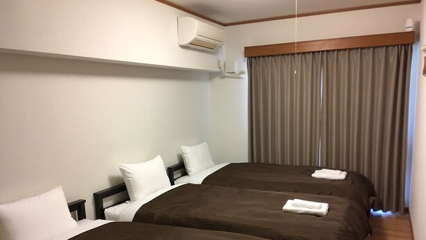 Une chambre d’hôtel avec deux lits, chacun recouvert d’une couverture marron et agrémenté d’oreillers blancs. On y trouve une fenêtre avec des rideaux, un climatiseur fixé au mur et un plancher en bois.
