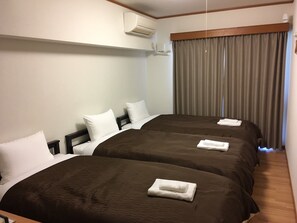 Habitación de hotel con dos camas, cada una con cubrecama marrón y almohadas blancas. Hay una ventana con cortinas, un equipo de aire acondicionado en la pared y piso de madera.