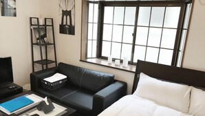 A hotel room with a black leather sofa, a bed with white bedding, a small table with a remote control, a blue folder, and a decorative shelf with books and a vase. - Tokyo Urban Flat Hotel 201 (Tokyo)