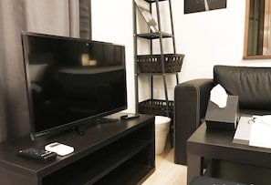 A modern living room with a flat-screen TV, a remote control, and a smartphone on a black entertainment unit. There is a black leather sofa and a small table with a tissue box and a ring box. A black metal shelf with baskets is against the wall. - Tokyo Urban Flat Hotel 201 (Tokyo)