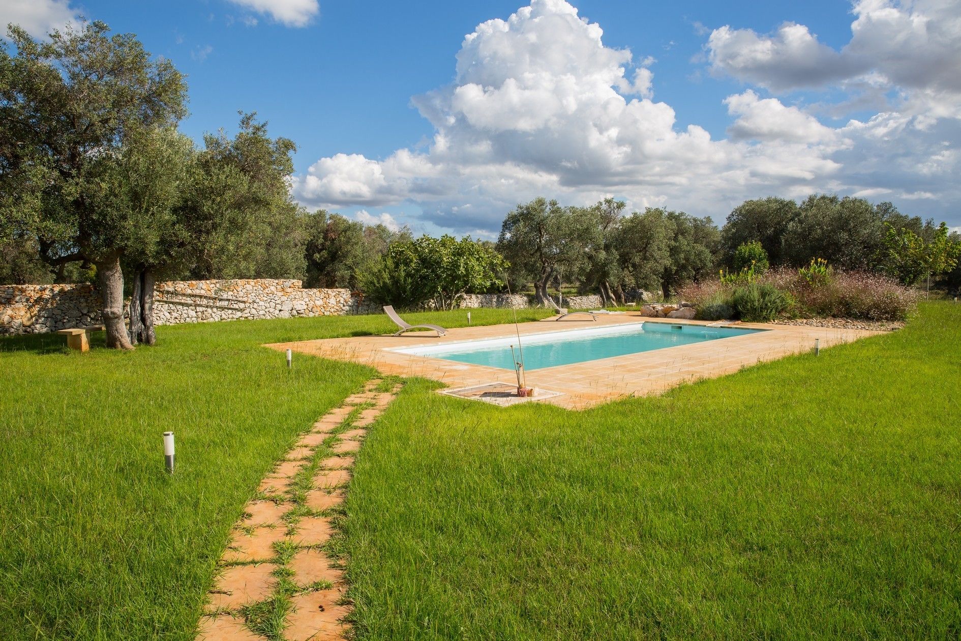 Une piscine entourée d’un mur de pierre, avec une pelouse et des arbres en arrière-plan.