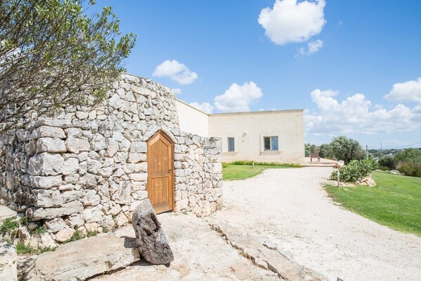 Un edificio de piedra con puerta de madera, un camino de entrada pavimentado y un edificio moderno al fondo.