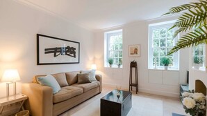 A modern living room with a beige sofa, a black coffee table, and a large window with a view of greenery. - Bright and Leafy 1 Bedroom Flat in the Heart of Chelsea (London)
