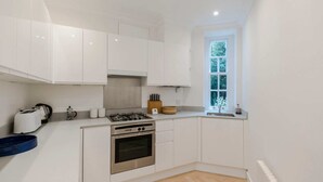 A compact kitchen with white cabinets, a gas stove, an oven, and a window with a view of greenery. - Bright and Leafy 1 Bedroom Flat in the Heart of Chelsea (London)