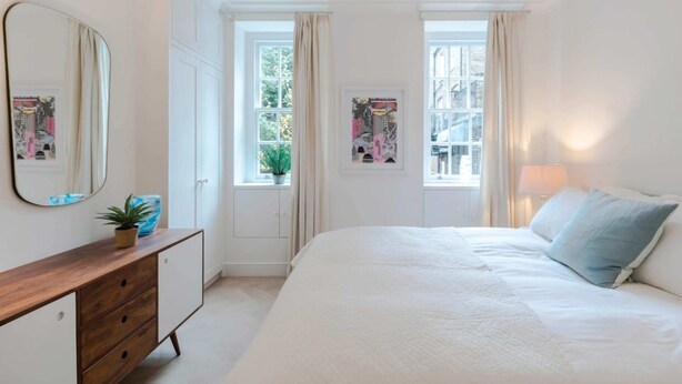 A bedroom with a large bed, a dresser, a mirror, and two windows with curtains. - Bright and Leafy 1 Bedroom Flat in the Heart of Chelsea (London)