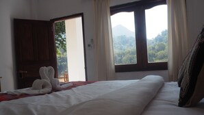 Quarto de hotel com uma cama, porta de madeira, janela com cortinas e vista para uma montanha.