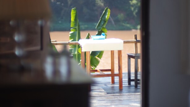 Vista pela janela com uma mesa e cadeiras de madeira, toalha de mesa branca e um fundo de vegetação exuberante.