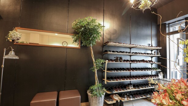 A shoe display with multiple shelves, a potted plant, and a bench in a room with dark walls and a chandelier. - Trip & Sleep Hostel (Nagoya)