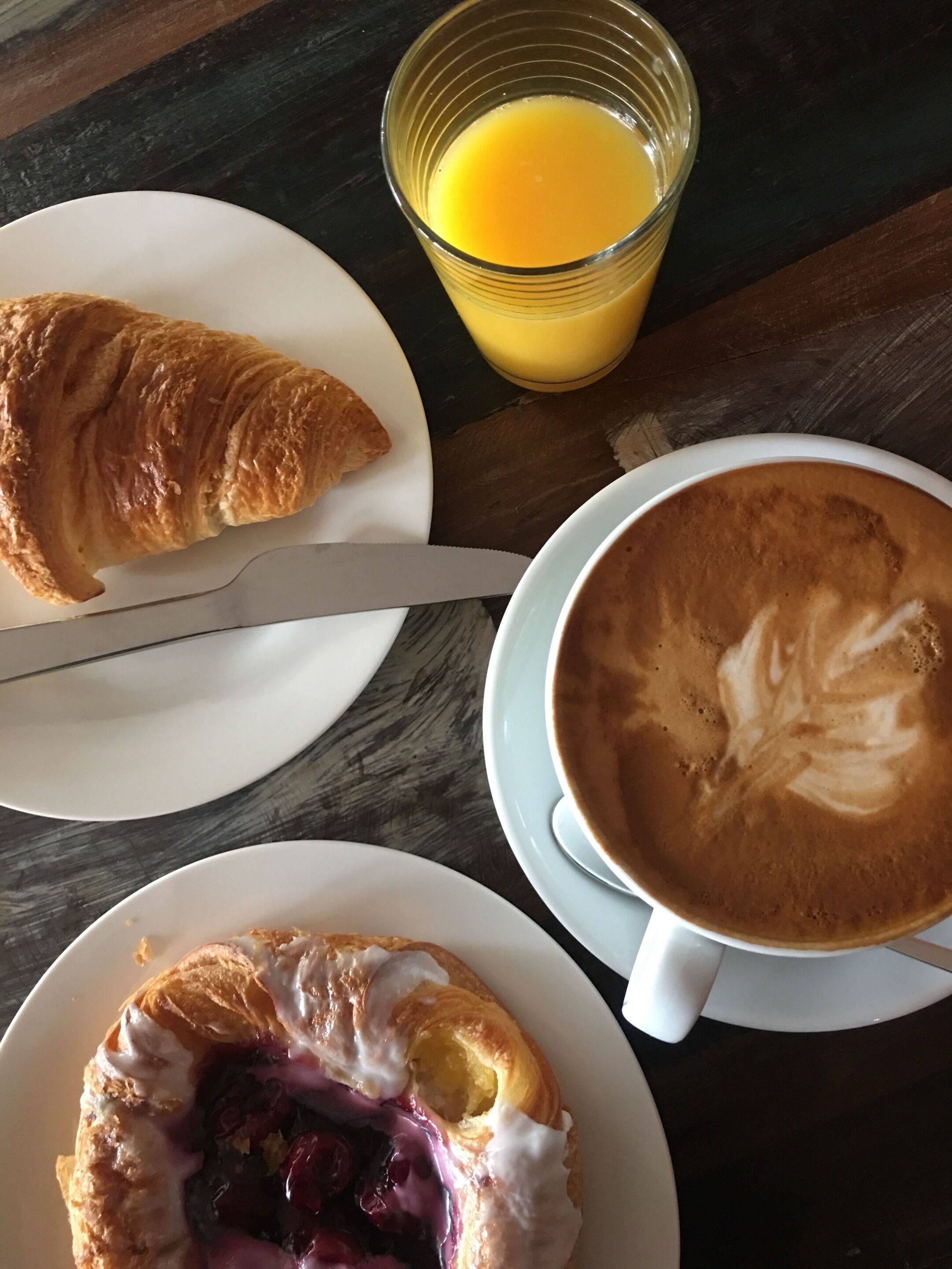 A breakfast setting with a croissant, a glass of orange juice, and a cup of coffee with latte art on a wooden table.