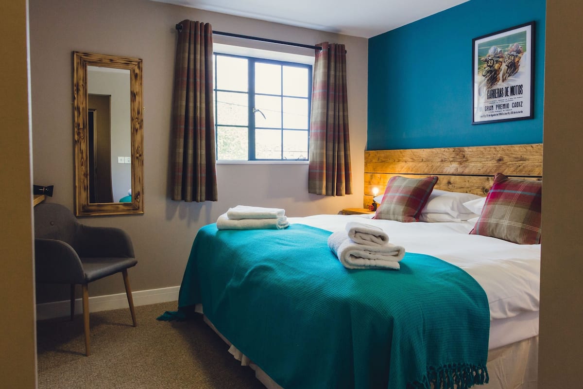 A hotel room with a bed, a chair, a window with curtains, a mirror, and a wall with a framed picture.