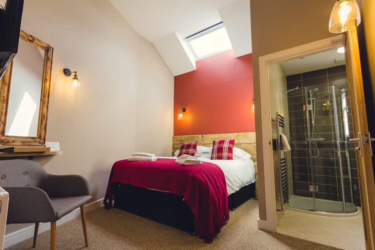 A hotel room with a bed, a chair, a mirror, a red wall, and a shower area.