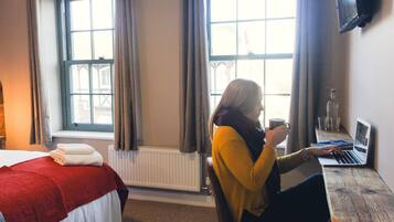 A hotel room with a bed, a desk with a laptop, and a window with curtains.
