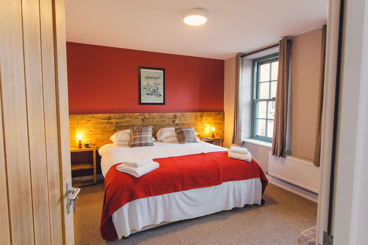 A bedroom with a large bed, a wooden headboard, a red wall, a window with curtains, and a framed picture on the wall.
