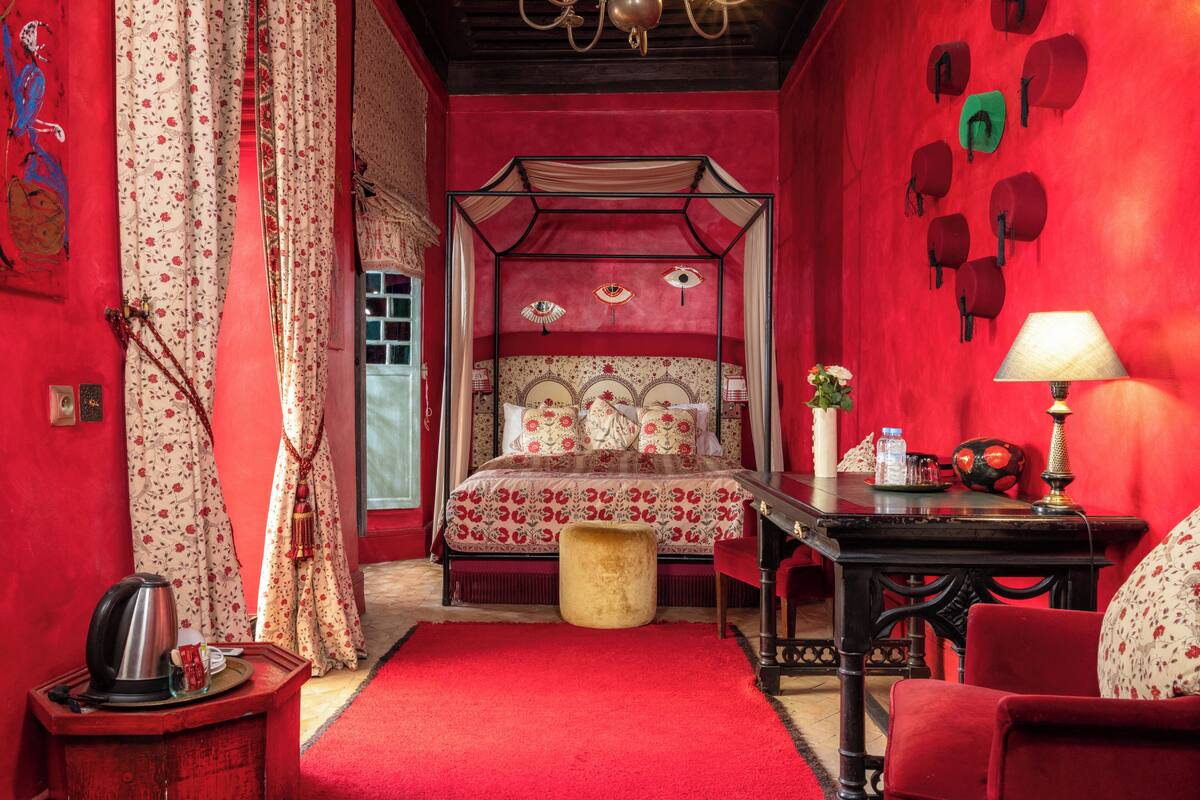 A red-themed bedroom with a canopy bed, a wooden nightstand, a red armchair, and a red carpet.
