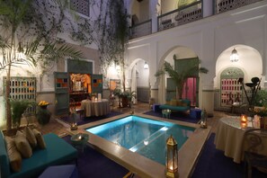 Un espace piscine intérieur luxueux, doté d’un coin salon et entouré d’arcades et de plantes.