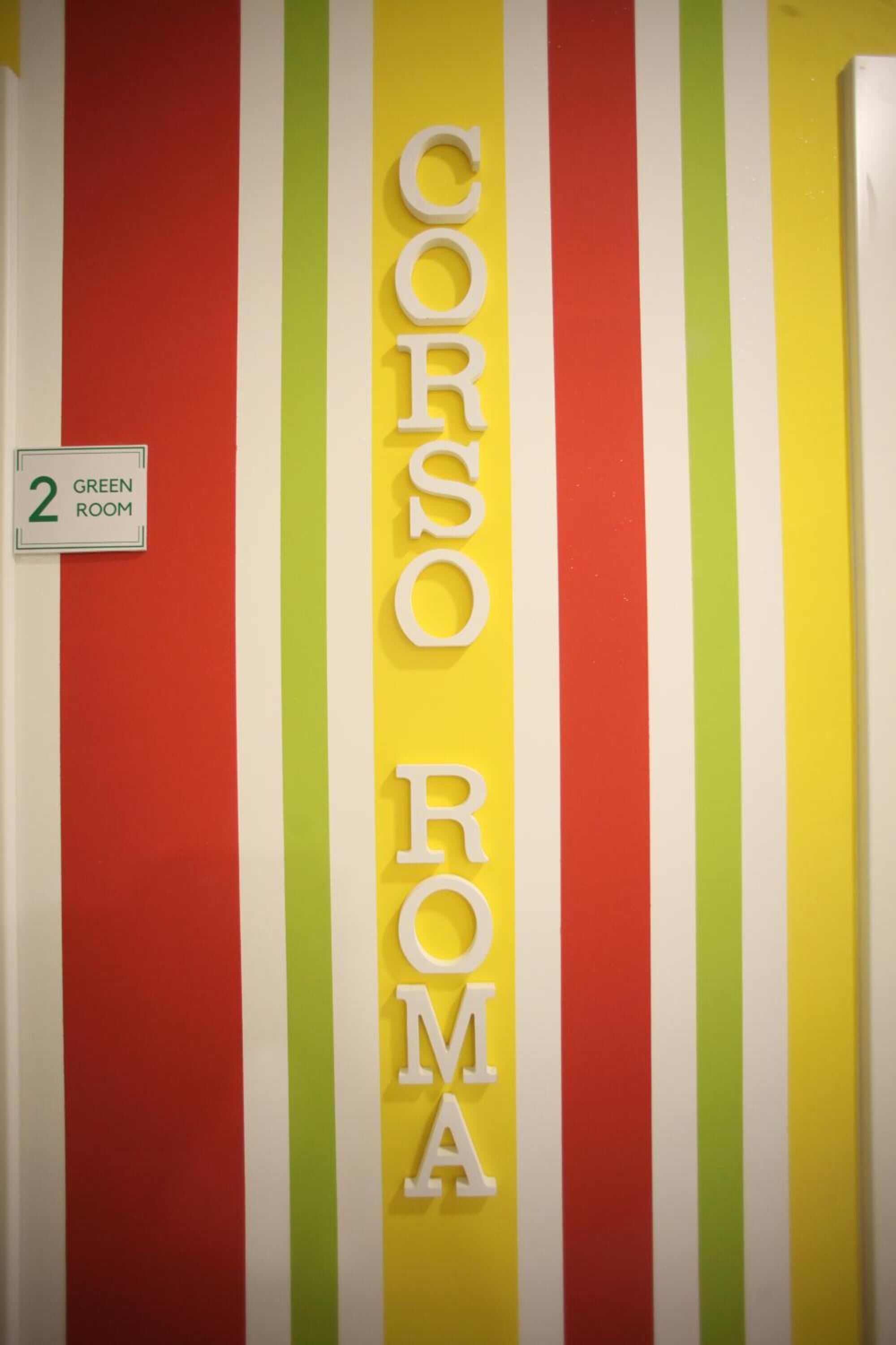 Un muro con strisce verticali rosse, gialle e verdi, con la scritta 'CORSO ROMA' in bianco. Un segnale indica 'GREEN ROOM' e il numero '2'.