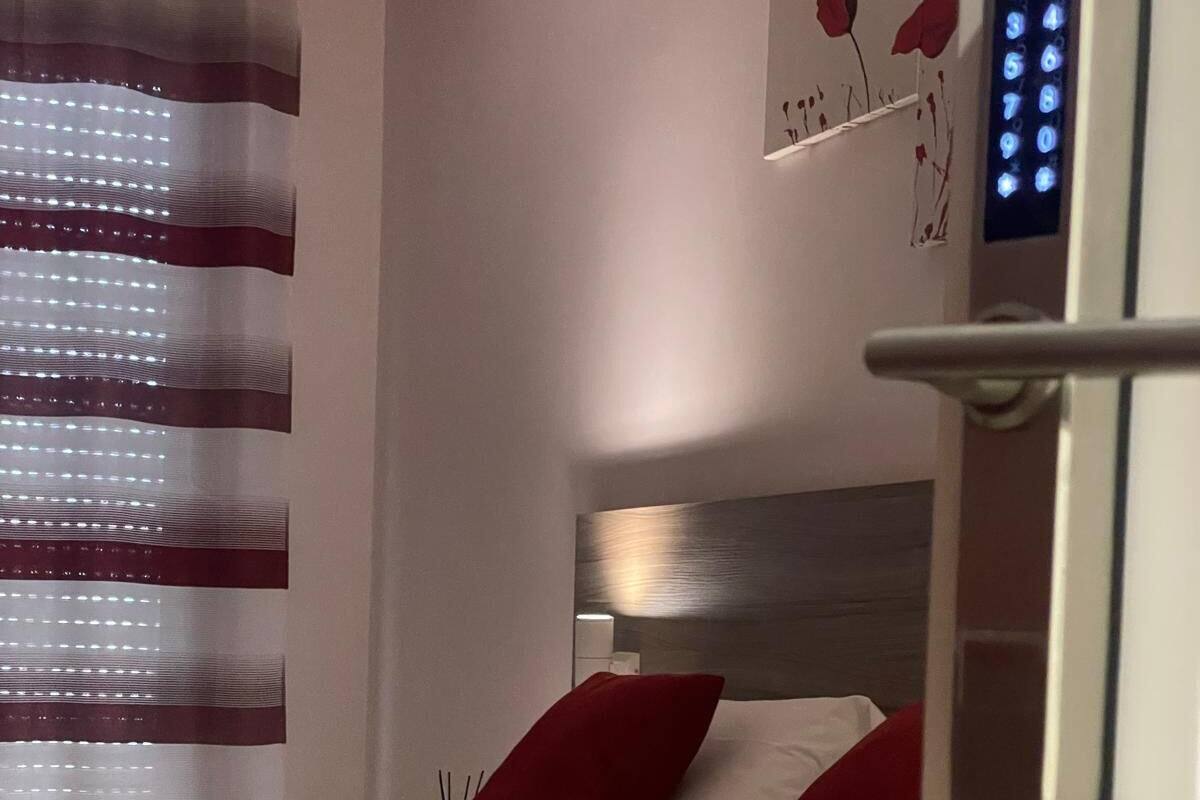 Une chambre d ’hôtel avec un lit, des oreillers rouges, une tête de lit, un panneau de commande fixé au mur et une fenêtre avec des stores.