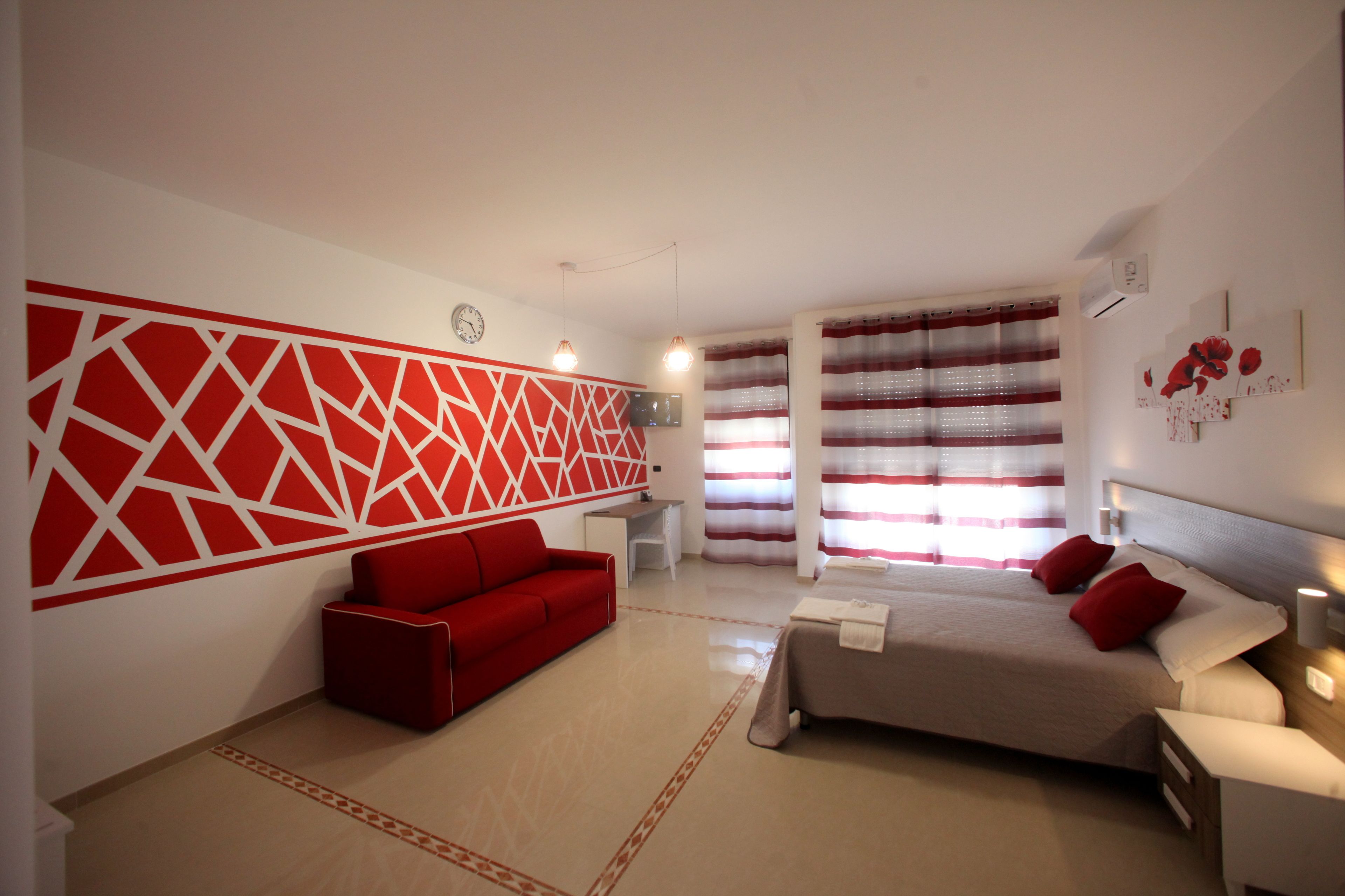 A modern hotel room with a red and white geometric wall design, a red sofa, a bed with red pillows, and a large window with striped curtains.