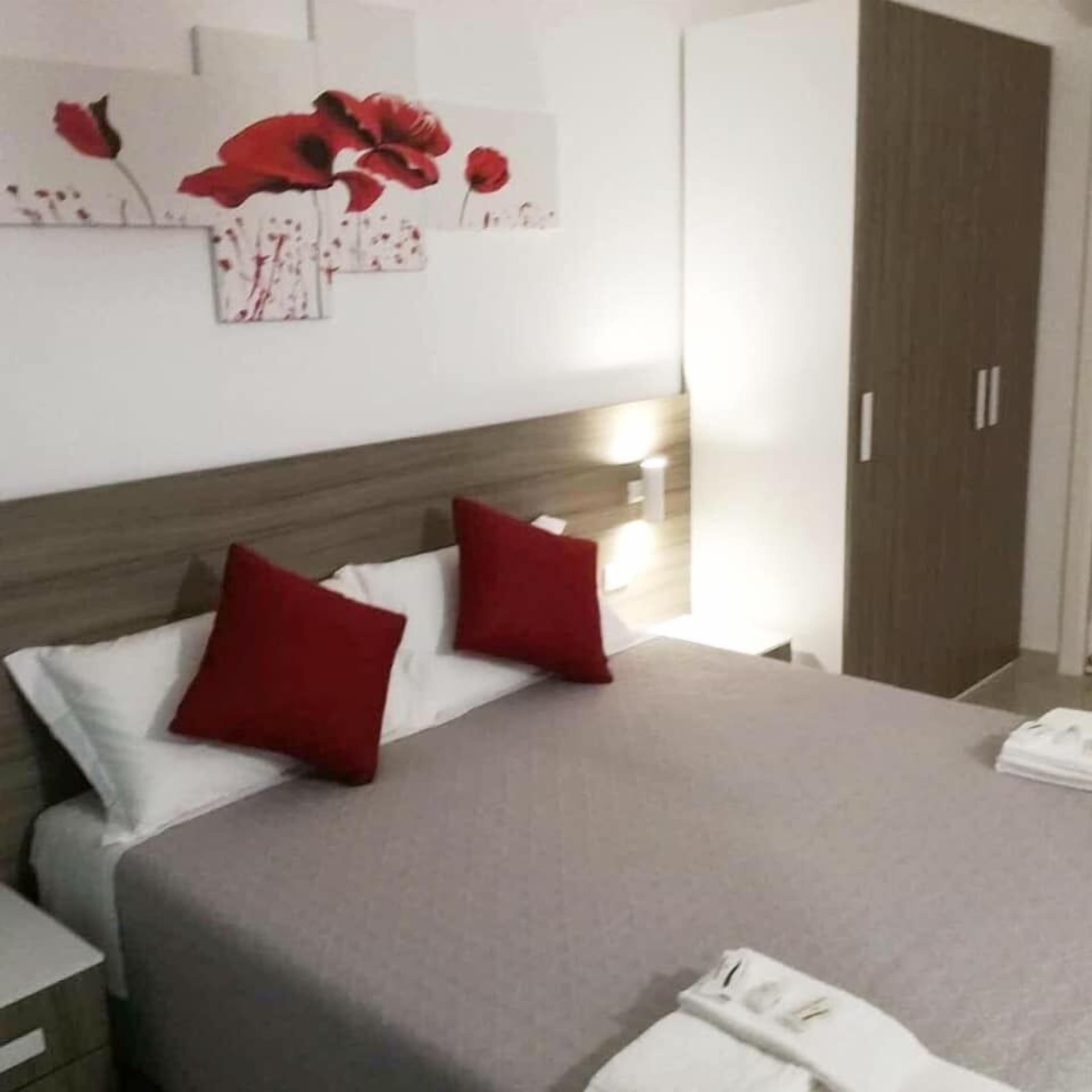 Una cama bien hecha con almohadas rojas y blancas, una colcha gris y un cabecero de madera. Se ve una mesita de noche con una lámpara y una toalla blanca doblada.