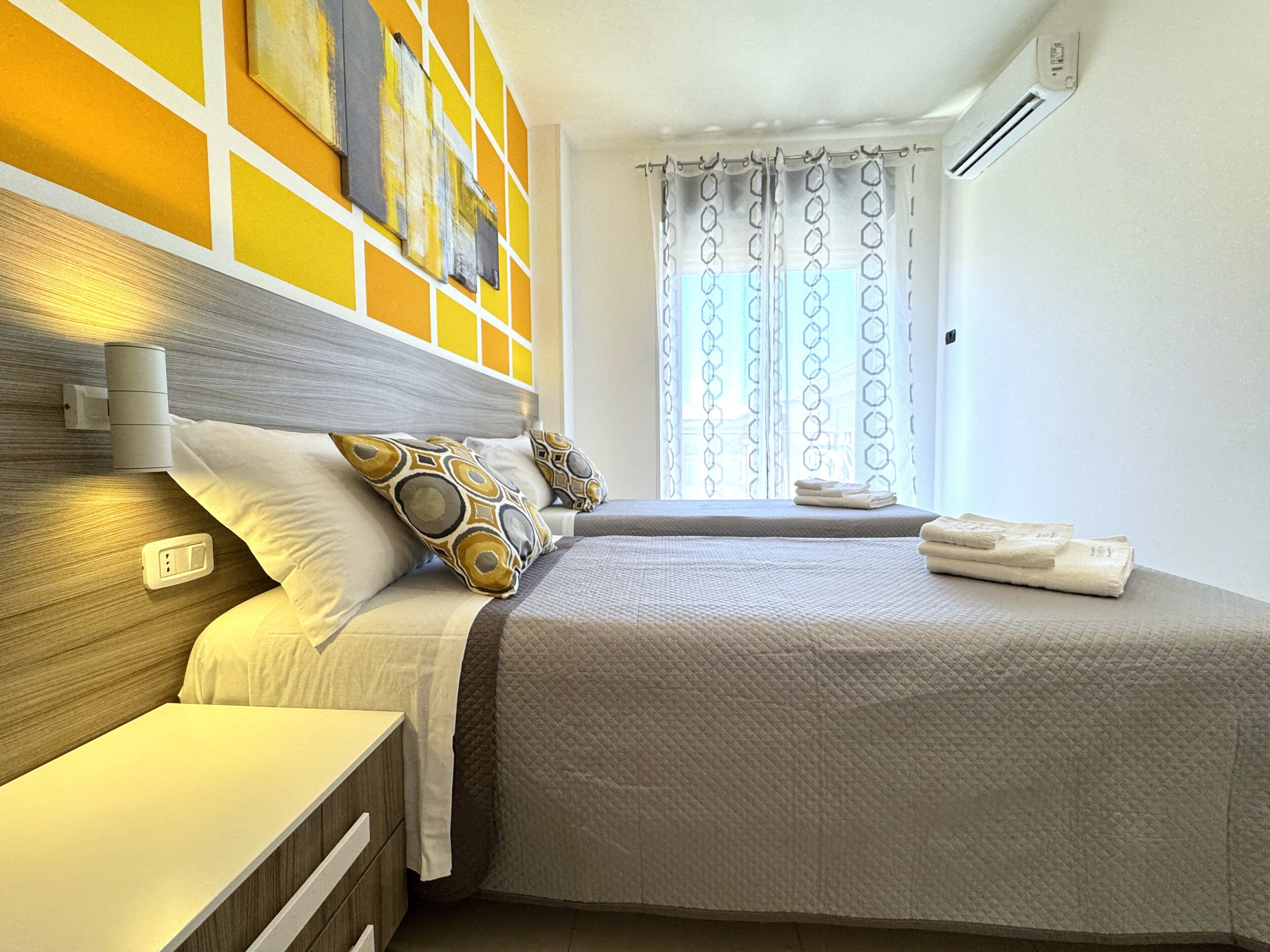 Habitación de hotel con cama, mesitas de noche, aire acondicionado instalado en la pared y ventana con cortinas estampadas.