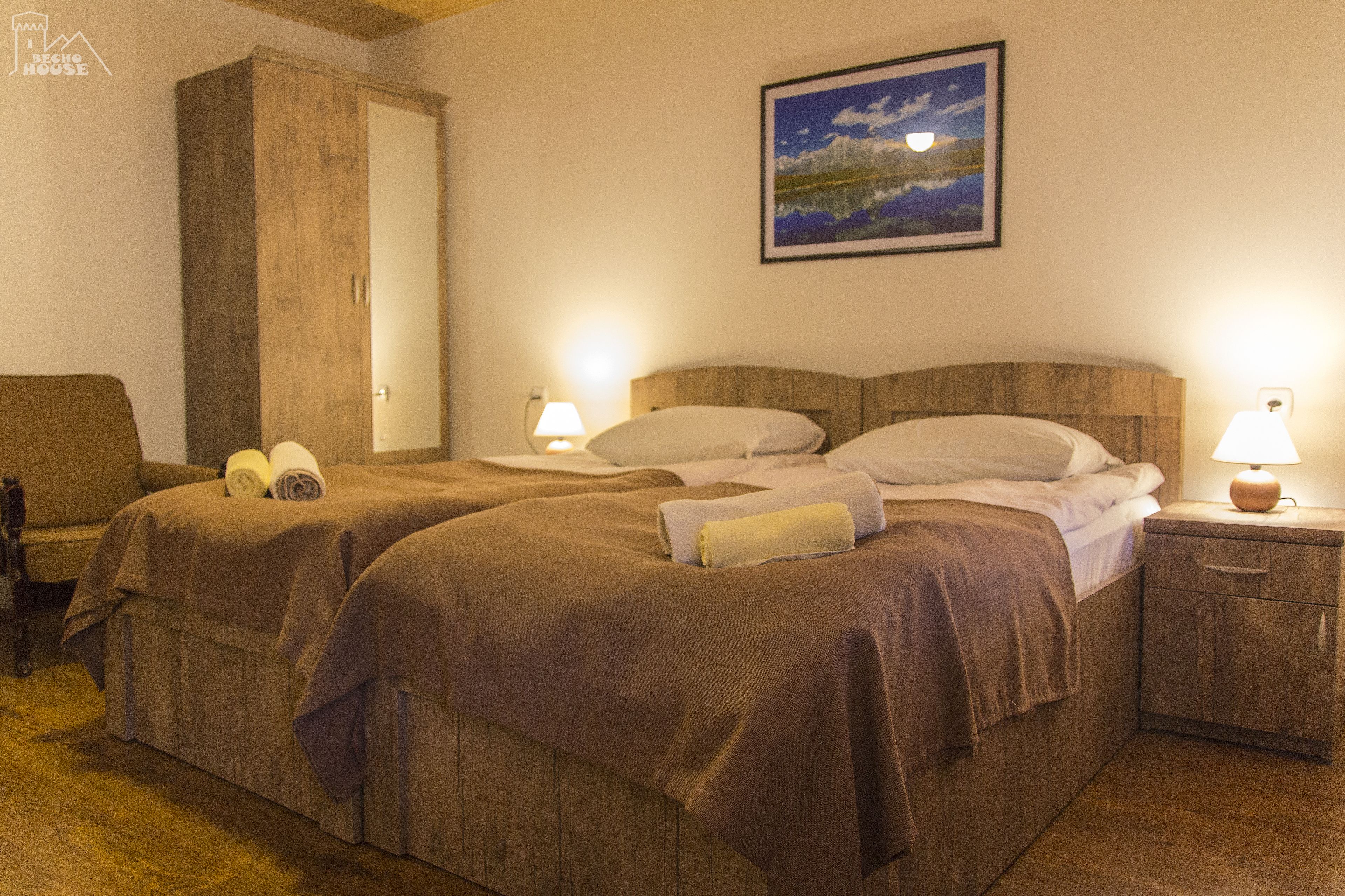Un dormitorio con dos camas, cabeceros de madera, una mesita de noche con lámpara, un armario y un cuadro en la pared.