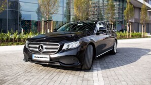 Un Mercedes-Benz negro estacionado frente a un edificio moderno con ventanas de vidrio y un área pavimentada.