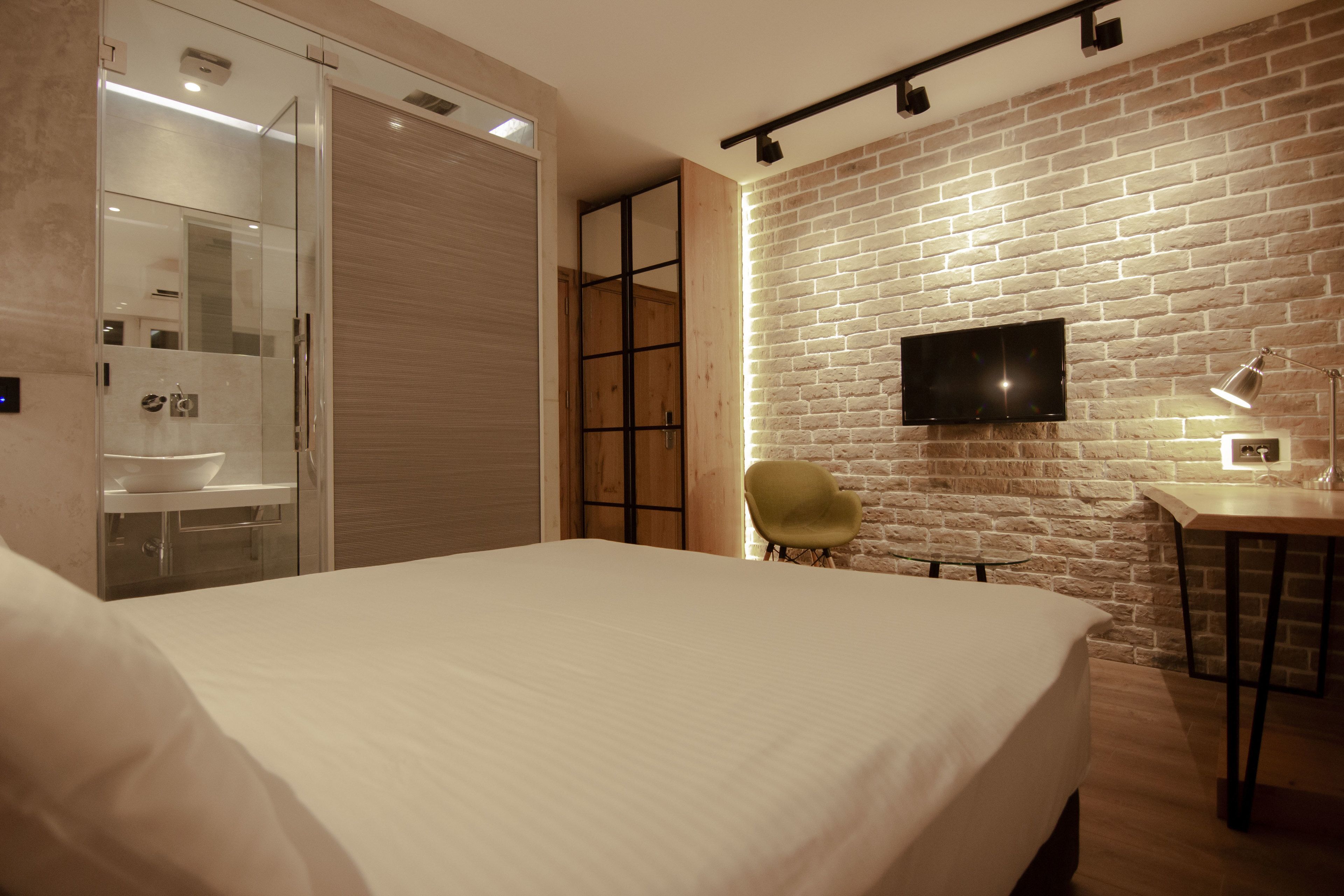 A hotel room with a large bed, a TV mounted on a brick wall, a desk with a lamp, and a chair.