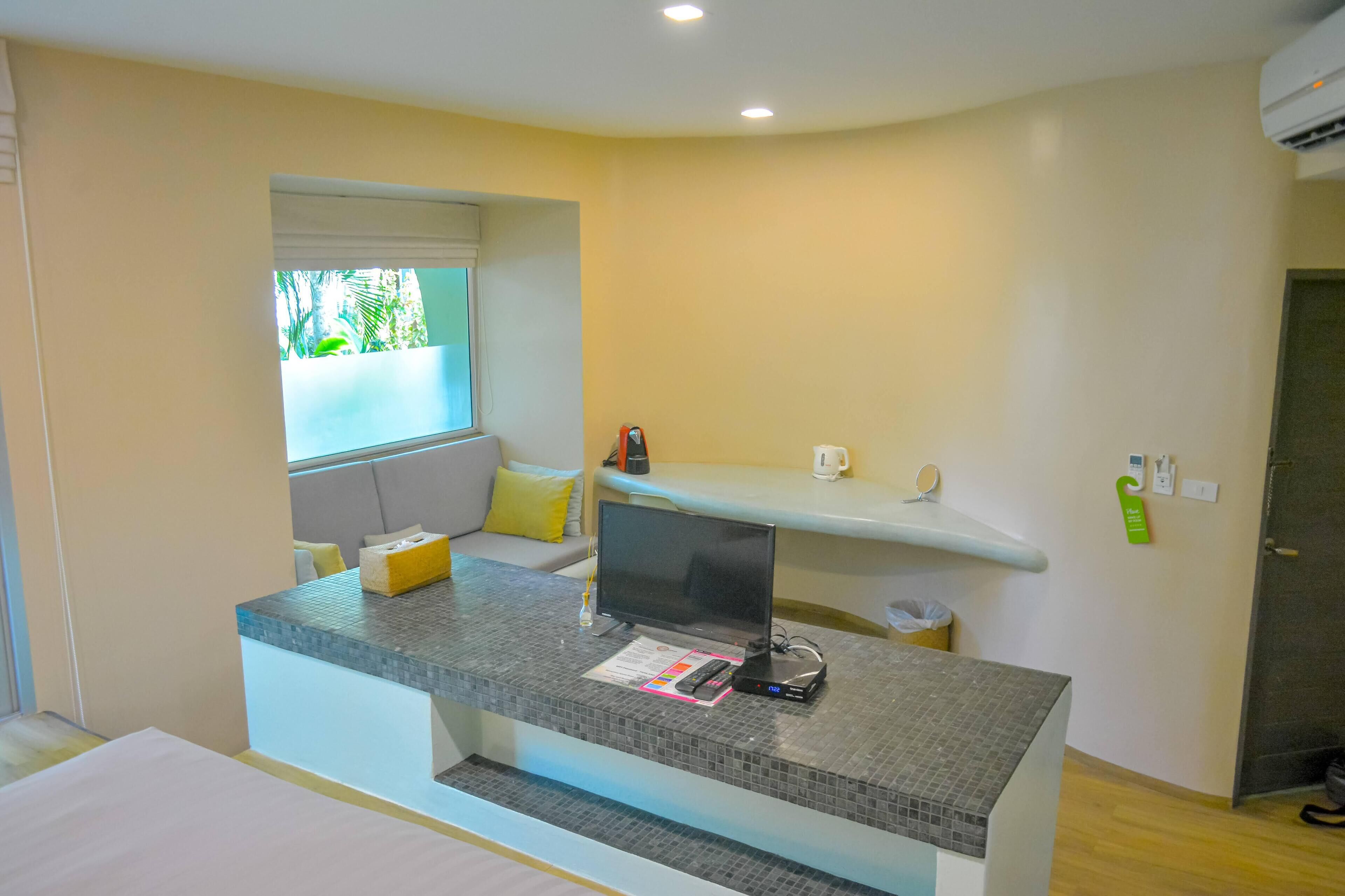 A modern hotel room with a grey tiled desk, a flat-screen TV, a small seating area with cushions, and a window with a view of greenery.