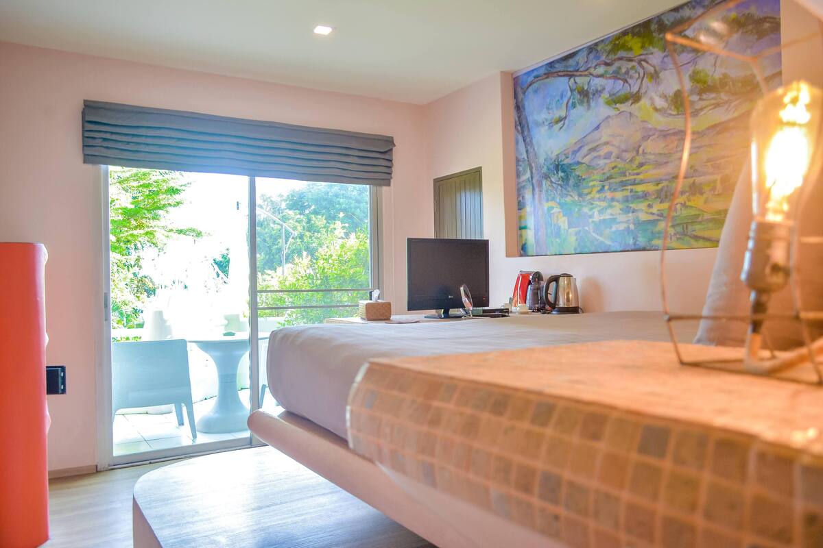 Un dormitorio con cama, televisor, un cuadro y ventana con vista a la vegetación.