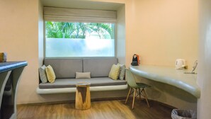 Una habitación moderna con sofá, un taburete de madera, una silla, ventana con vistas a zonas verdes y una kitchenette con tetera y cafetera.