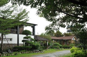 A traditional Japanese-style building with a wooden exterior and a tiled roof, surrounded by lush greenery and a gravel path.