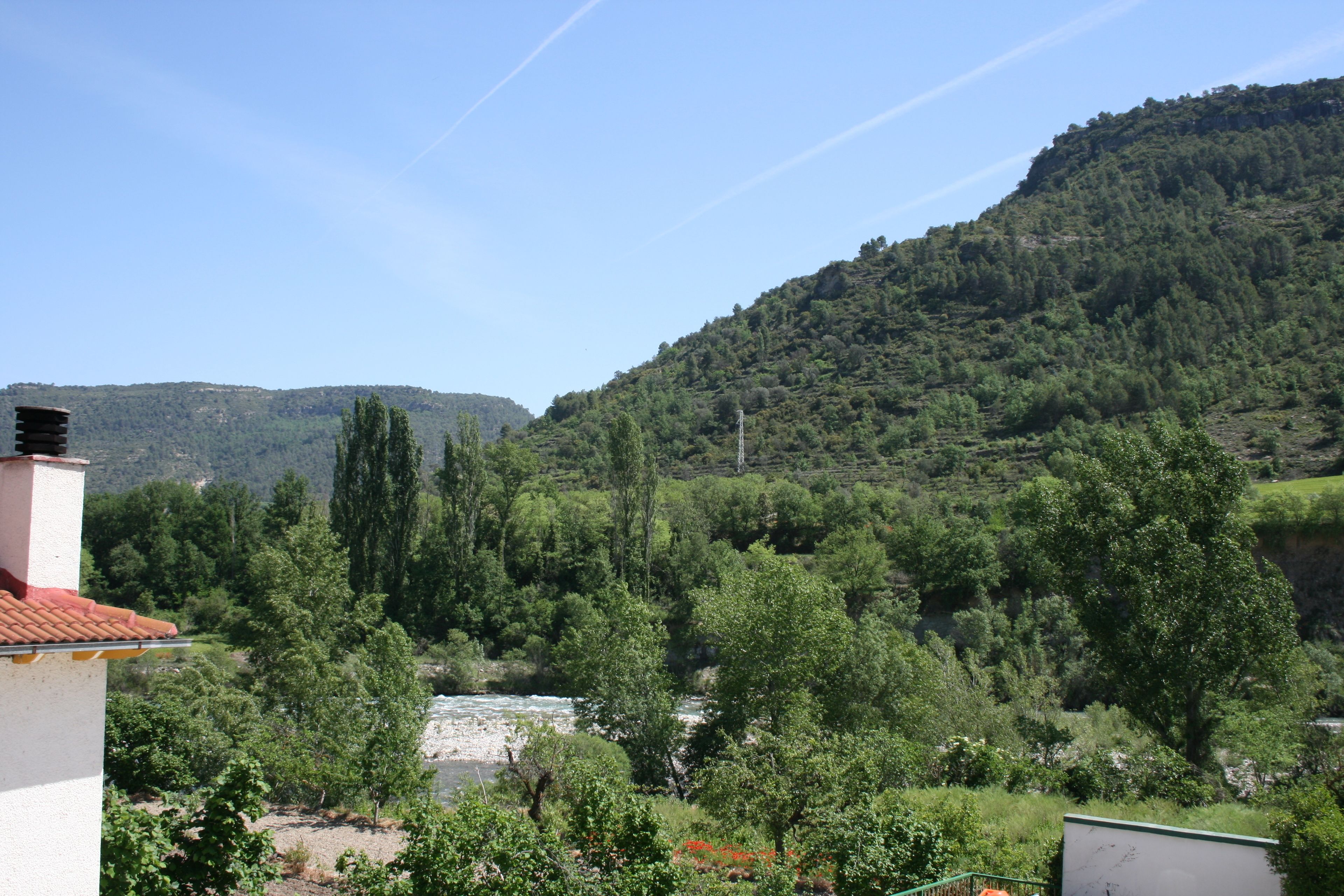 Une vue sur une rivière, avec des montagnes en arrière-plan, le tout entouré de verdure et agrémenté d’un bâtiment dont le toit est recouvert de tuiles rouges.