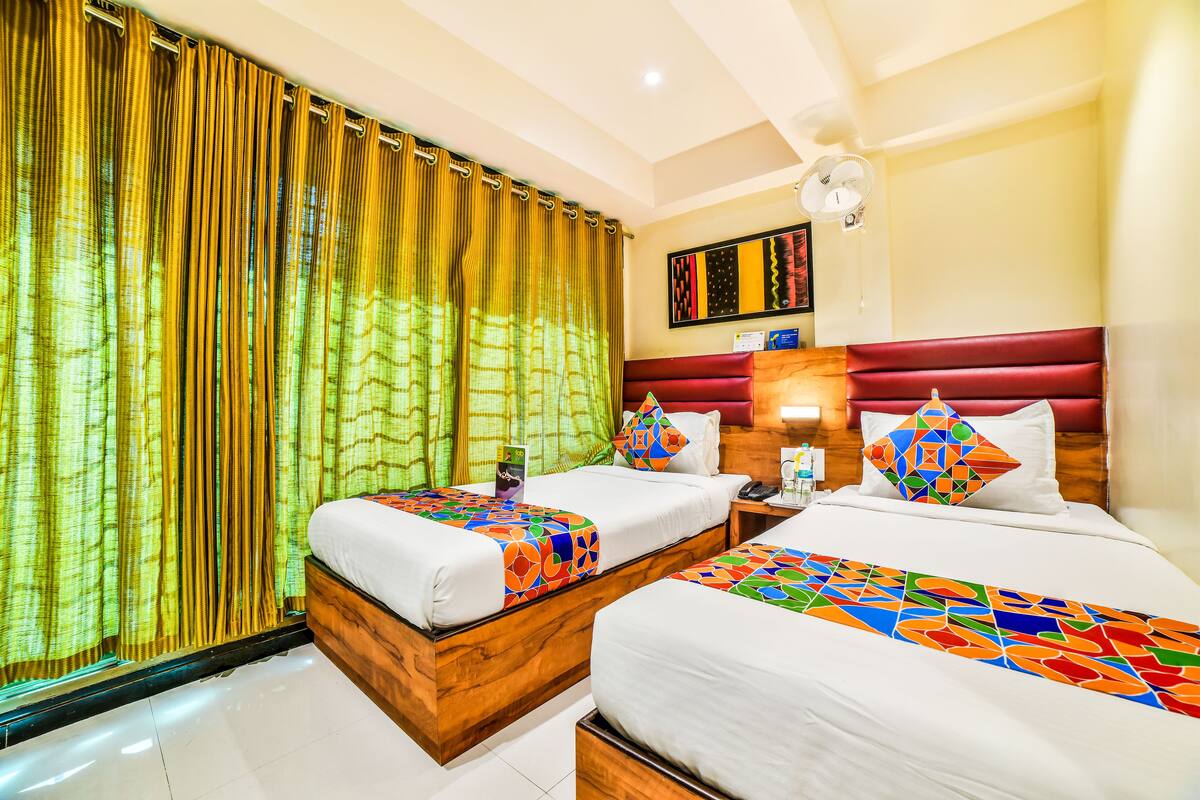 A hotel room with two beds, a wooden headboard, a bedside table with a lamp, and a window with curtains.