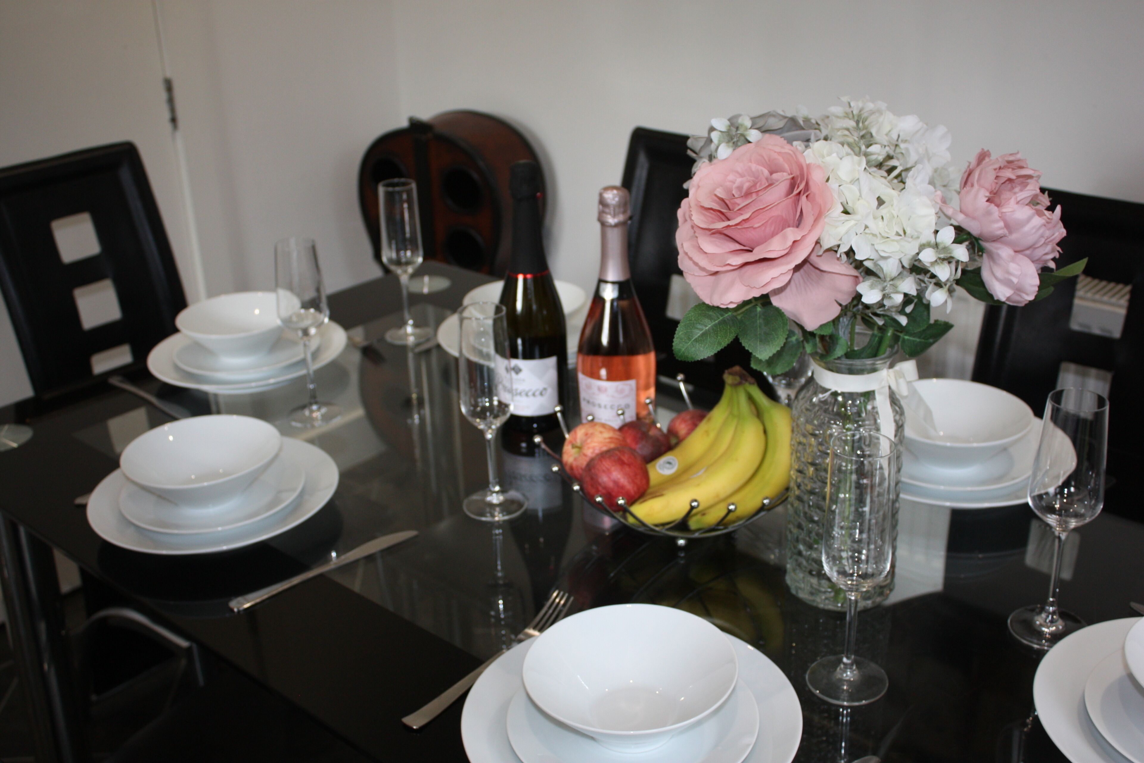 A dining table set with plates, bowls, wine glasses, a bottle of wine, a vase with flowers, and a fruit bowl.