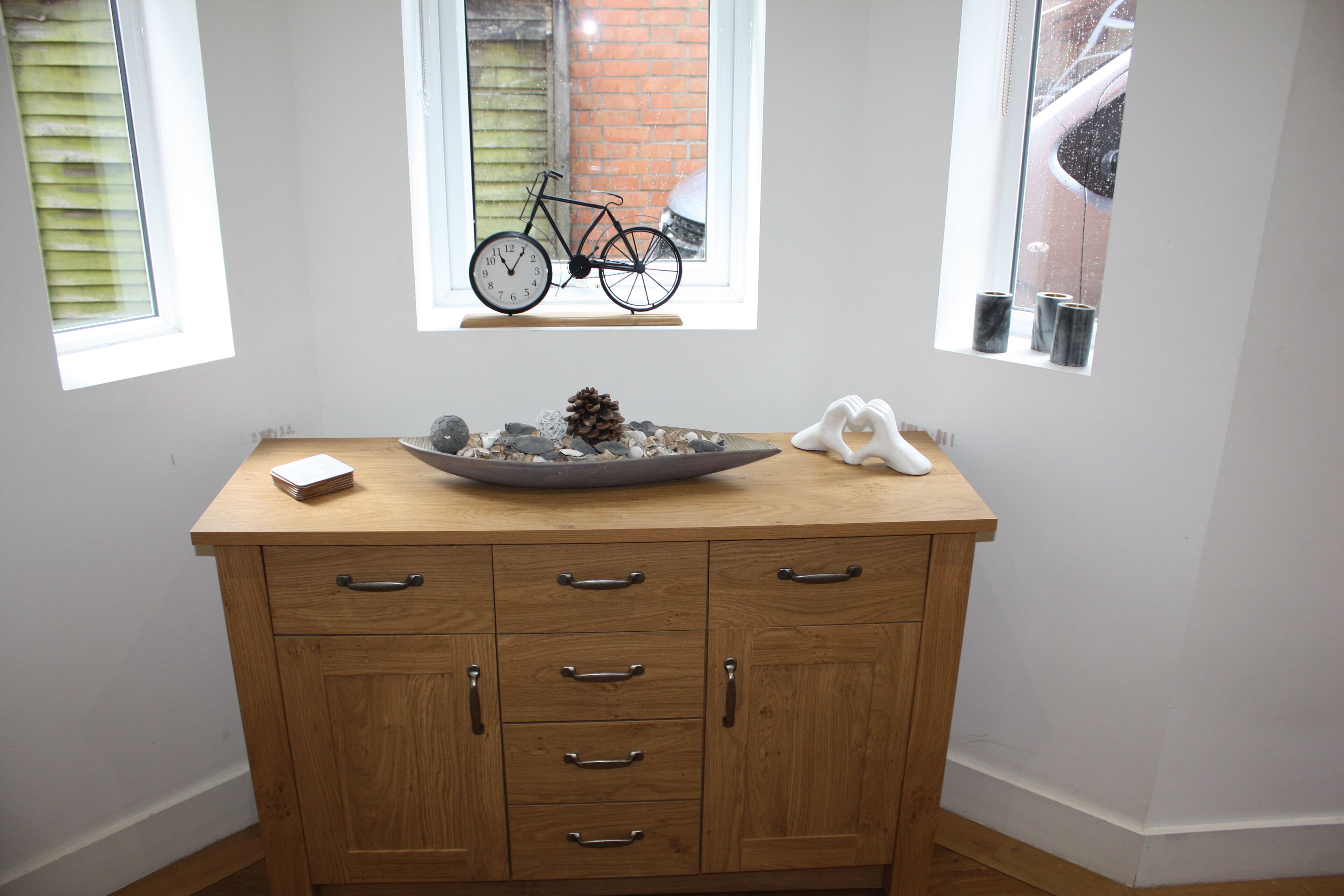 A wooden cabinet with drawers and a bowl on top, a window with a bicycle outside, and a small shelf with candles.