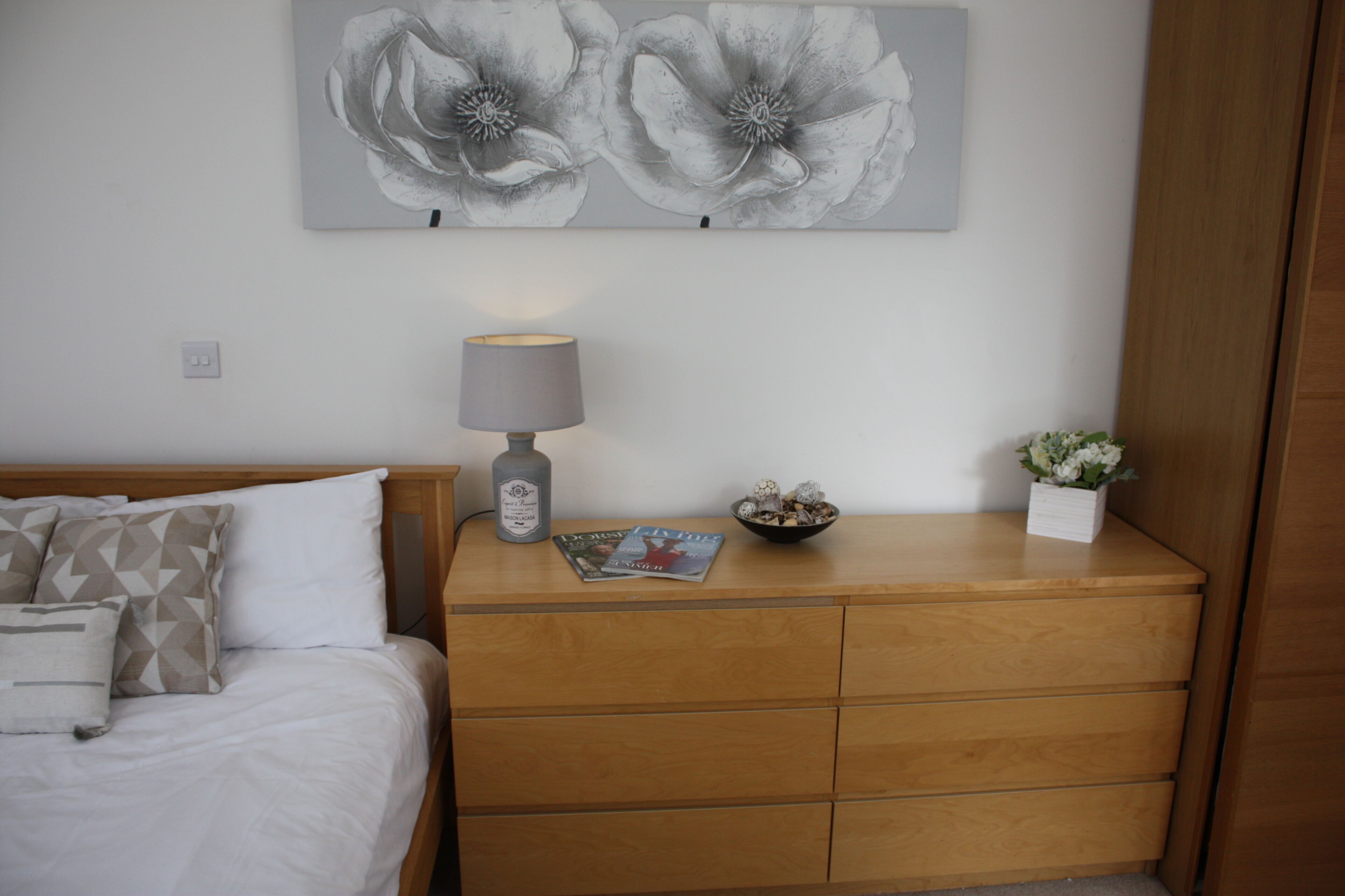 A neatly made bed with pillows, a bedside table with a lamp, a magazine, a decorative bowl, and a potted plant.