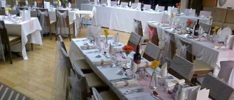 A banquet hall with multiple tables set for a formal event, featuring white tablecloths, silverware, and floral centerpieces.