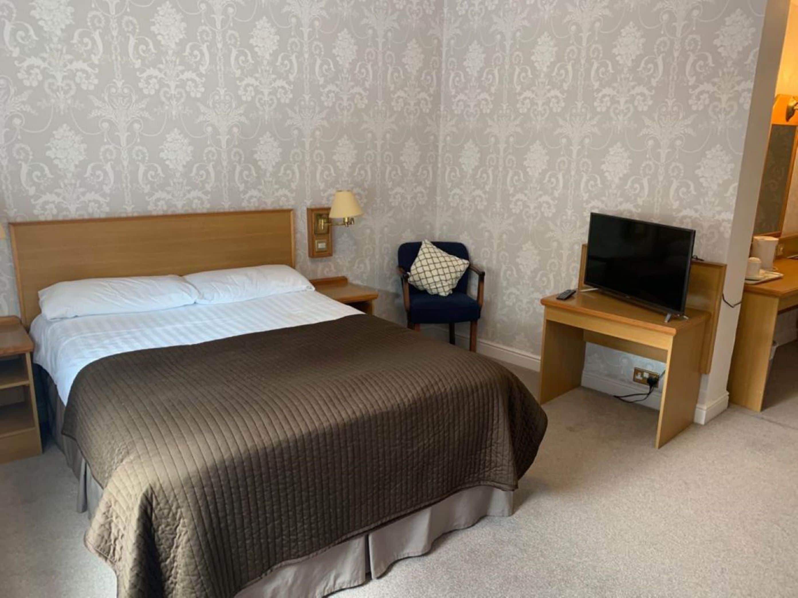 Habitación de hotel con una cama grande, un televisor sobre un soporte de madera, una silla y una mesita de noche con una lámpara.
