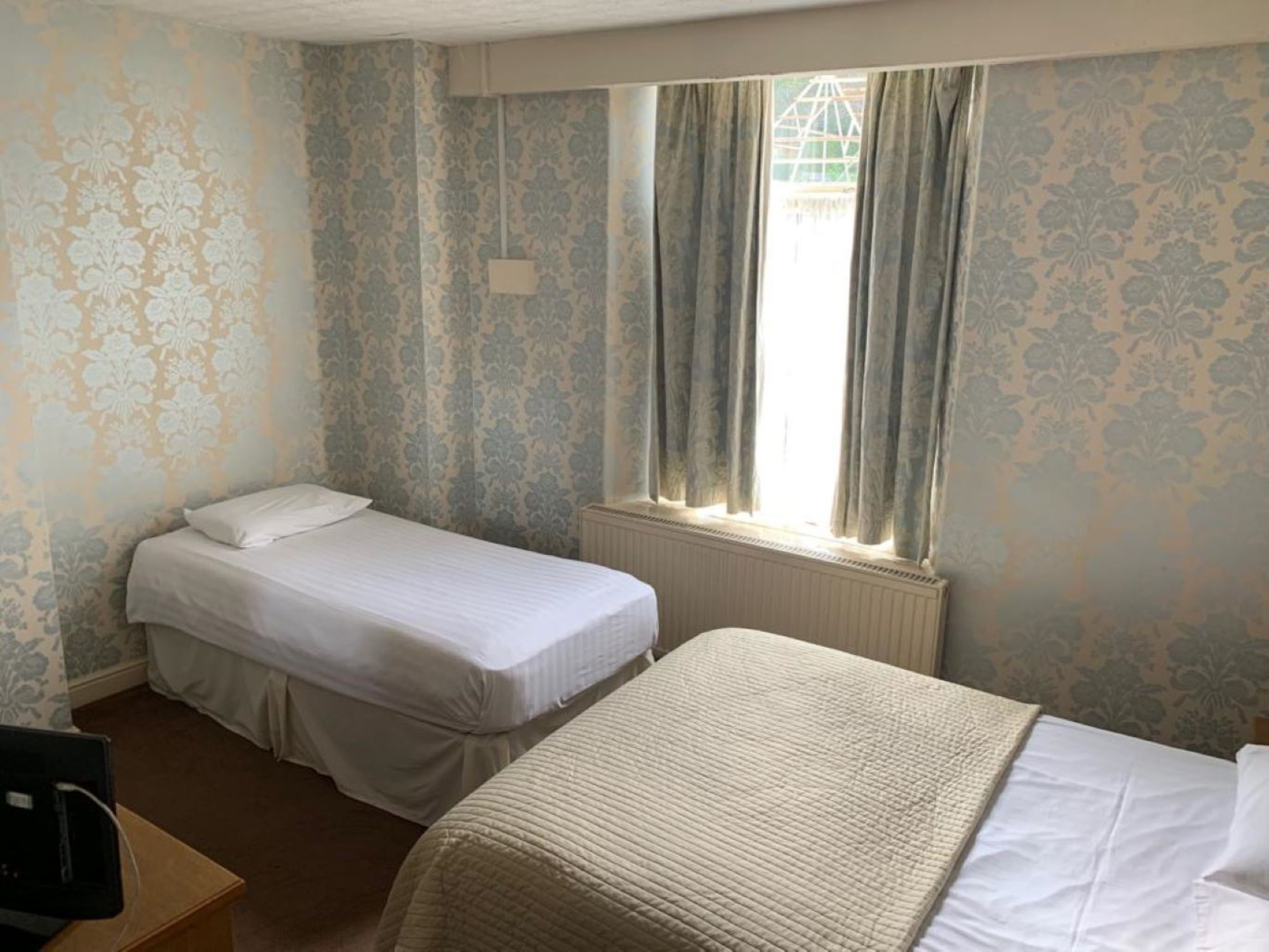 Ein Zimmer mit zwei Einzelbetten, einem Heizkörper, einem Fenster mit Vorhängen und Wandtapete mit Muster.