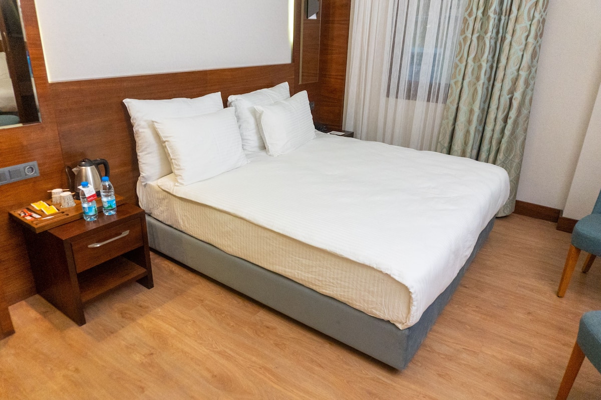 A hotel room with a large bed, wooden headboard, a nightstand with toiletries, and a chair.