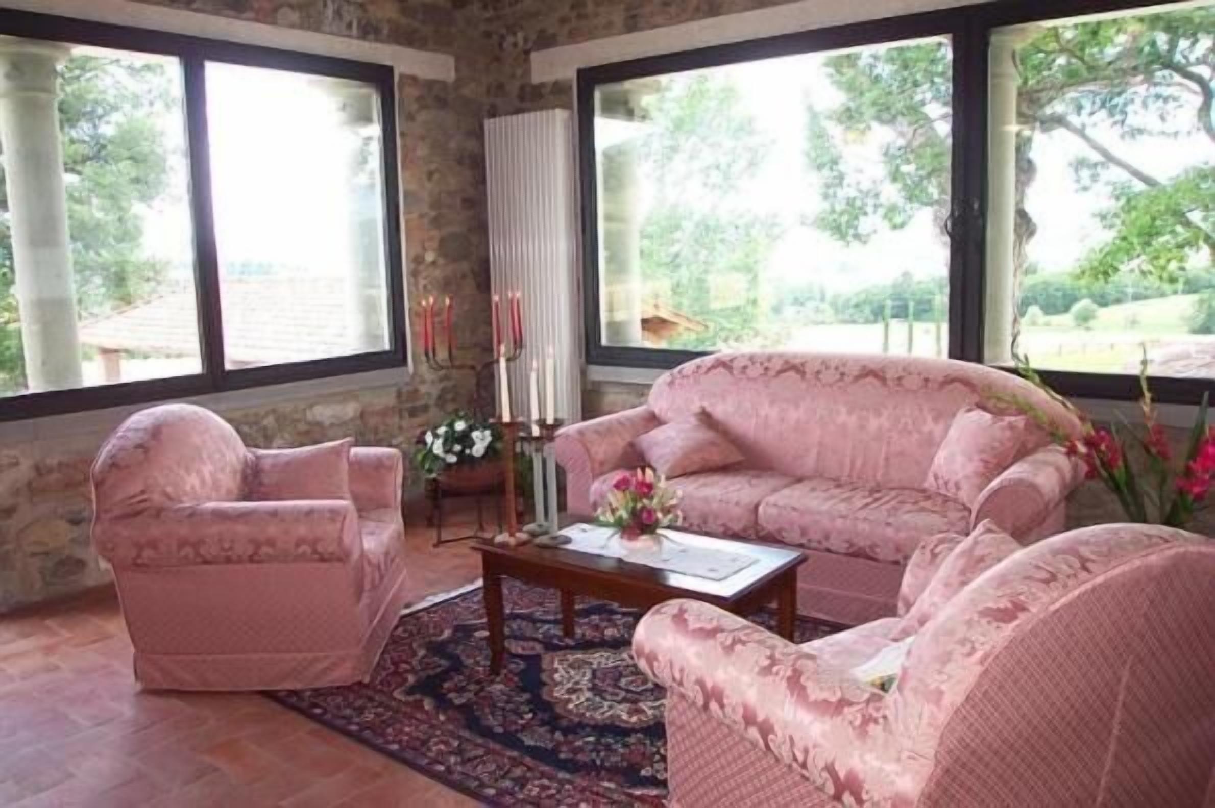 Una habitación con pared de piedra, ventanas amplias, un sofá rosa, un sillón a juego y una mesa de centro con un arreglo floral.