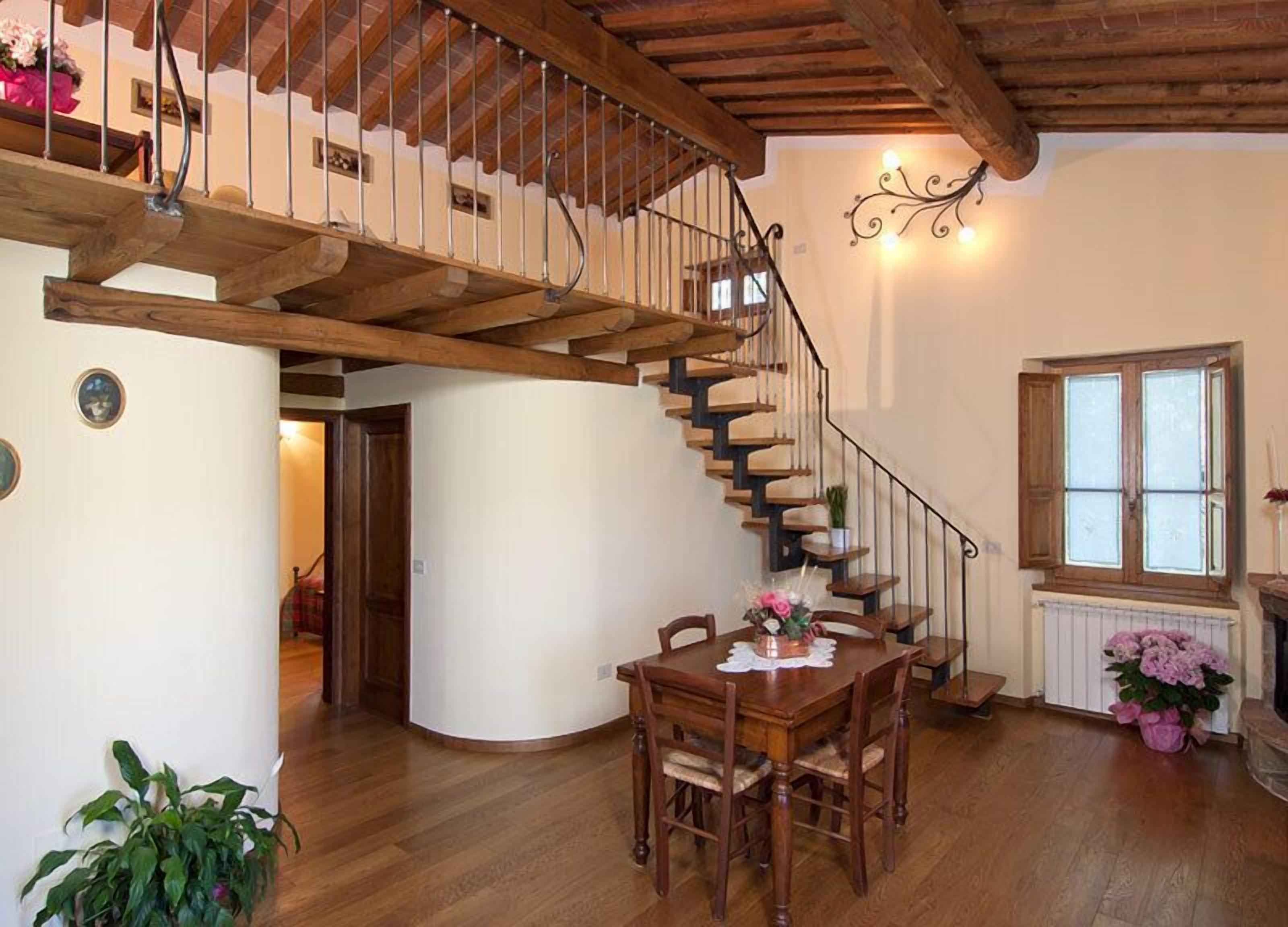 Una habitación amplia con una escalera de madera que conduce a un nivel superior, un comedor con mesa y sillas, y una ventana con vistas.