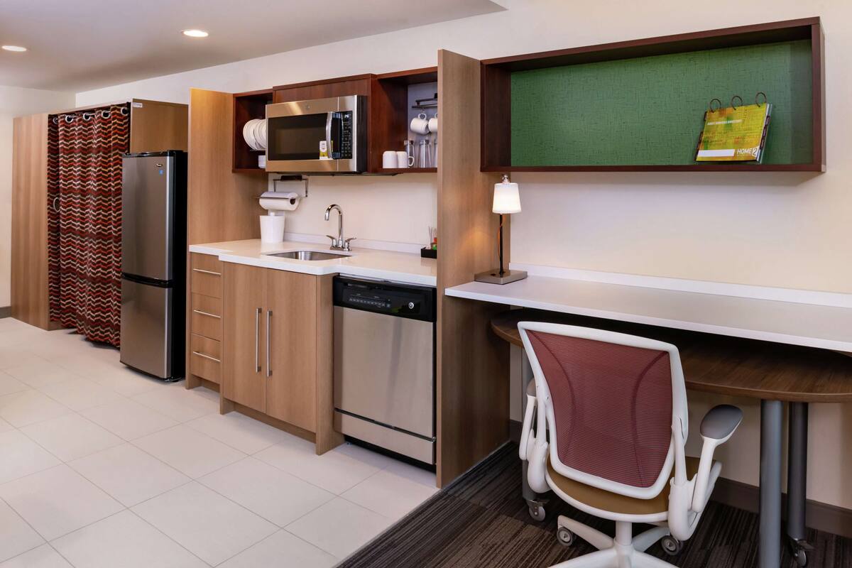 A modern kitchen with stainless steel appliances, wooden cabinets, a built-in microwave, and a small dining area with a white chair.
