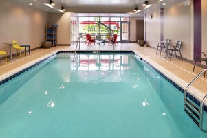 Ein Hallenbad mit klarem Wasser, umgeben von gelben und roten Stühlen, einem kleinen Tisch und einem Handtuchregal.