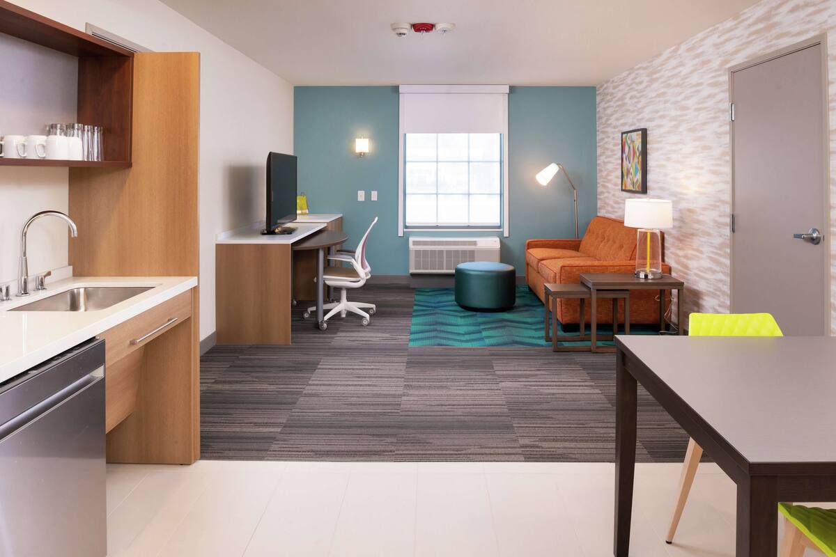 A compact hotel room with a kitchenette, a seating area with a sofa and armchair, a small table, and a window with blinds.
