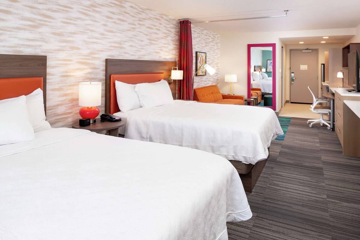 A hotel room with two beds, a desk, a chair, and a bathroom visible through an open door.