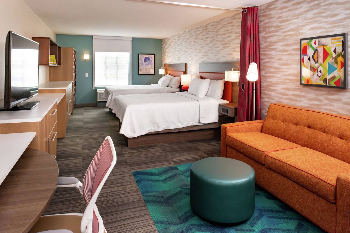 A hotel room with two beds, a desk with a computer, a TV, a sofa, and a patterned rug.
