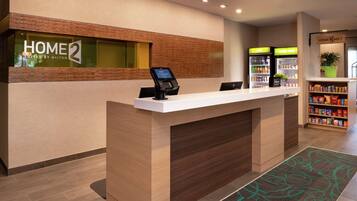 A hotel reception desk with a digital kiosk, a minibar, and a self-service beverage station.